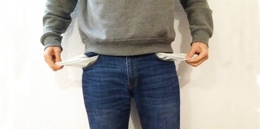 nome sujo - foto de um homem branco, vestido uma suéter cinza e uma calsa jeans, puxando os fundos dos bolsos vazios para fora.