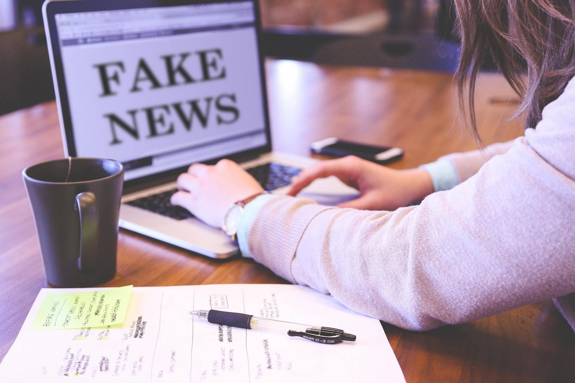 fake news e indenizacao - imagem de uma mulher branca, vestindo uma roupa executiva, sobre uma mesa de madeira, digitando as paavras fake news no notebook