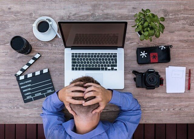 exclusão indevida de conta de rede social - foto de um homem branco, usando camisa social azul, com as mãos sobre a cabeça, apoiado em uma mesa que contém um laptop, uma câmera de vídeo, uma planta, uma xícara de café
